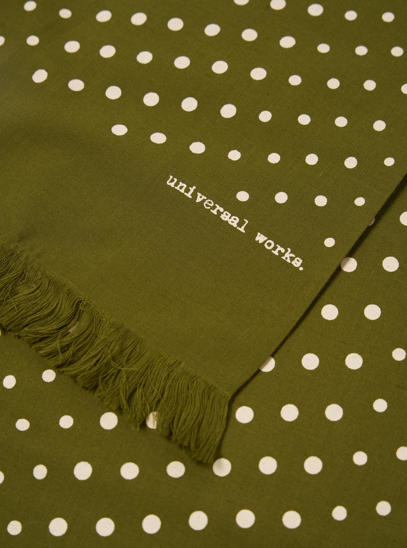 Close-up of a Green Dot Print Short Scarf. The olive green scarf has white polka dots and a fringed edge. The words "universal works." are printed on the fabric.
