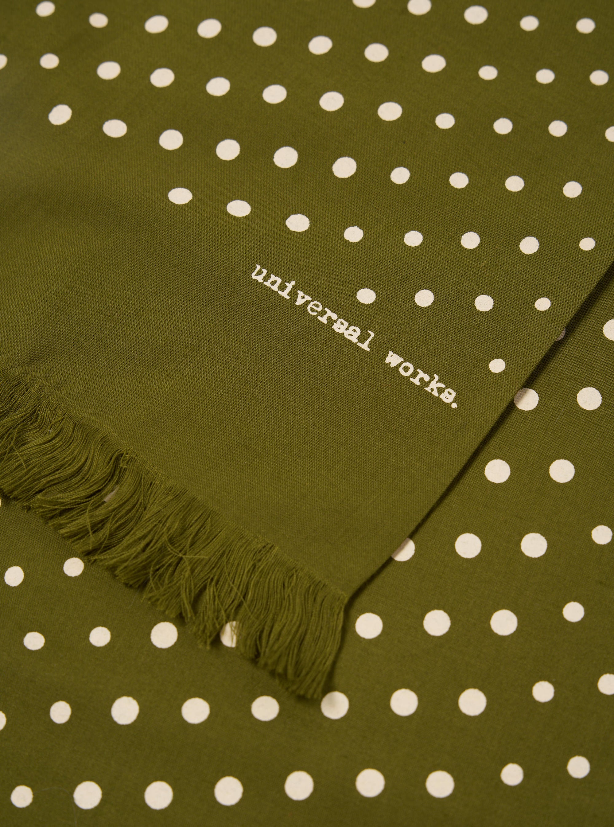 Close-up of a Green Dot Print Short Scarf. The olive green scarf has white polka dots and a fringed edge. The words "universal works." are printed on the fabric.
