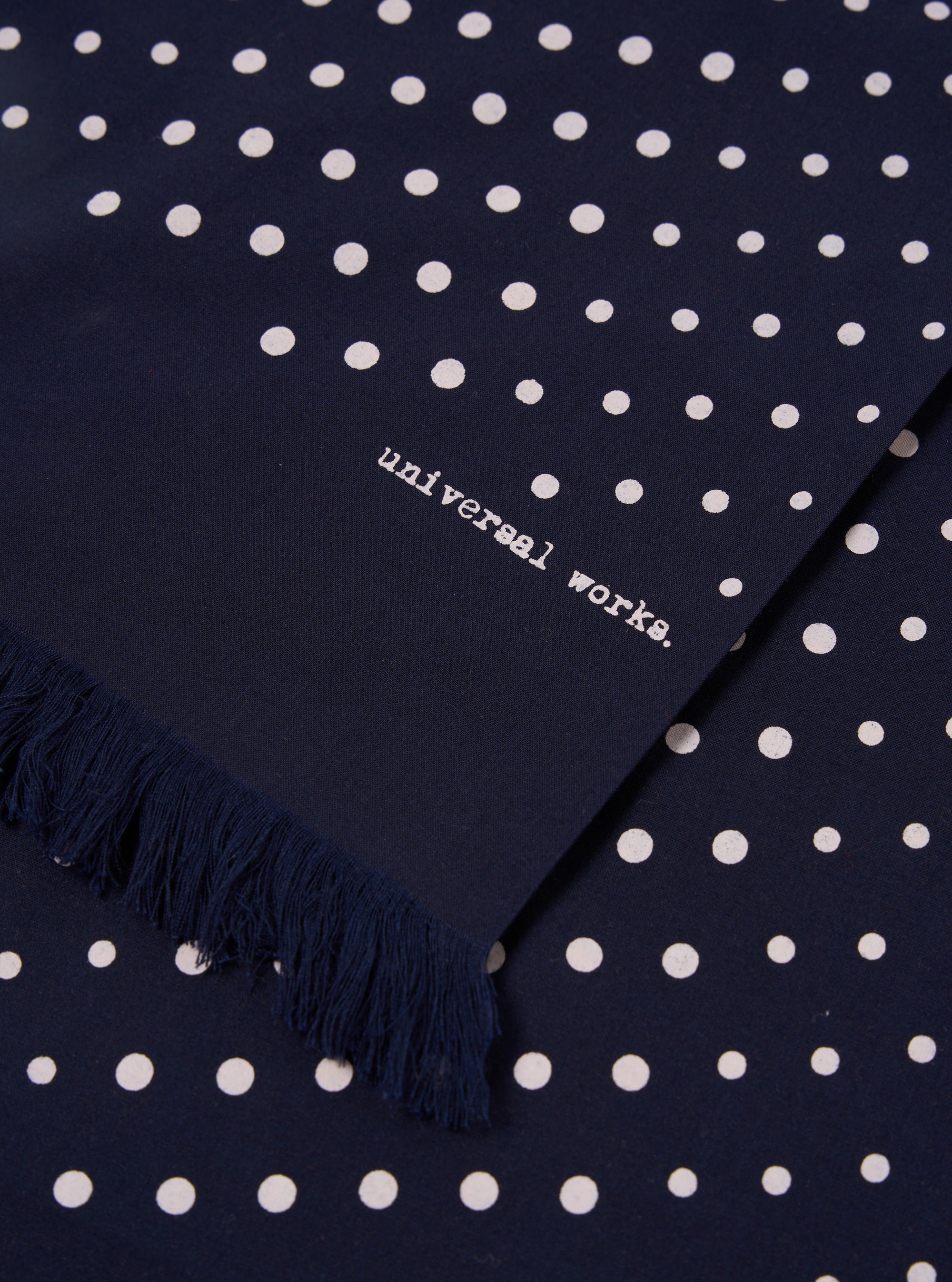 Close-up of a Navy Dot Print Short Scarf with fringe. The scarf is navy with white dots and the words "universal works." are printed on it.