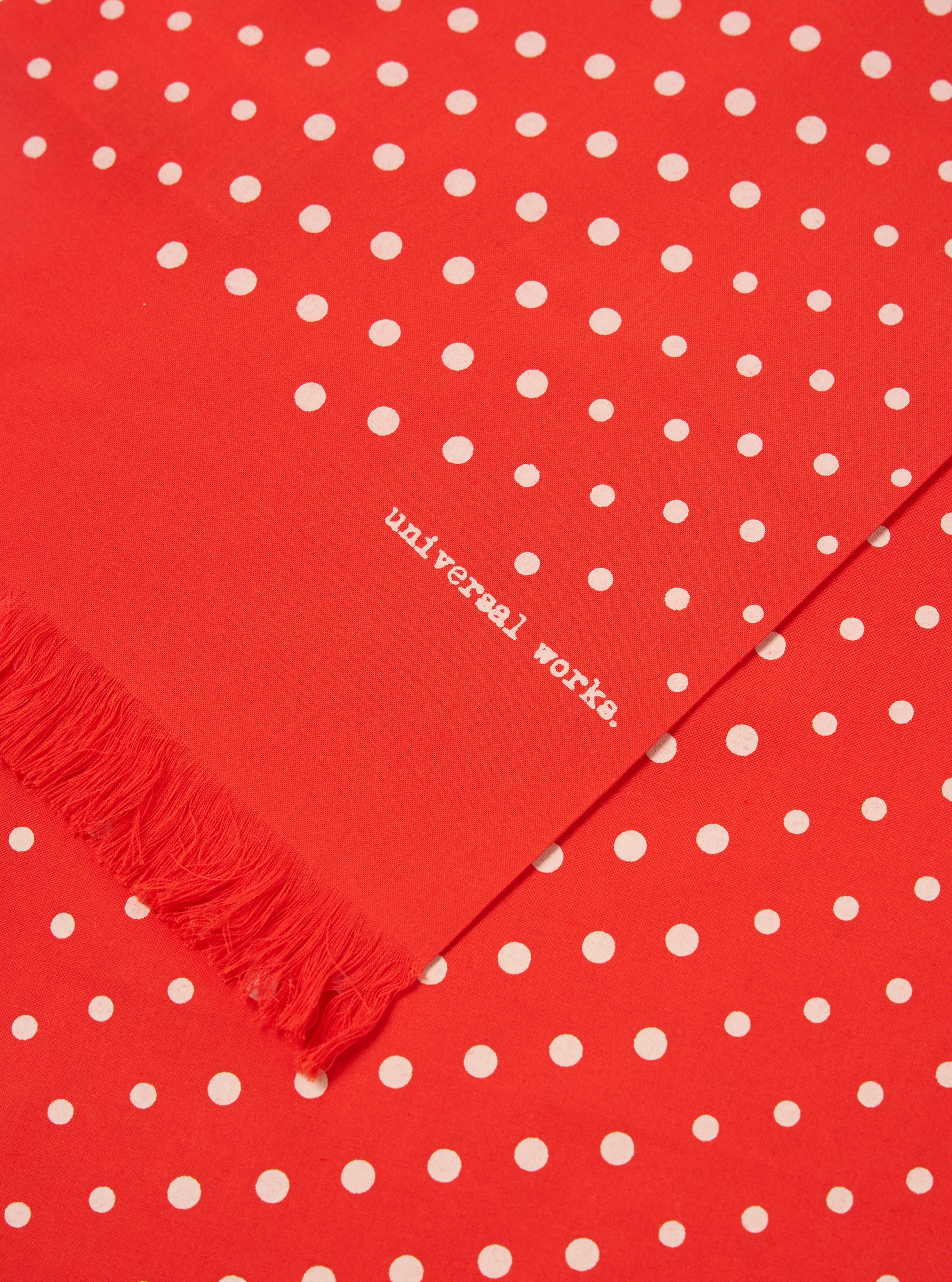 Close-up of an Orange Dot Print Short Scarf. The scarf is red with white polka dots and a fringed edge. The words 'universal works' are printed on the scarf.