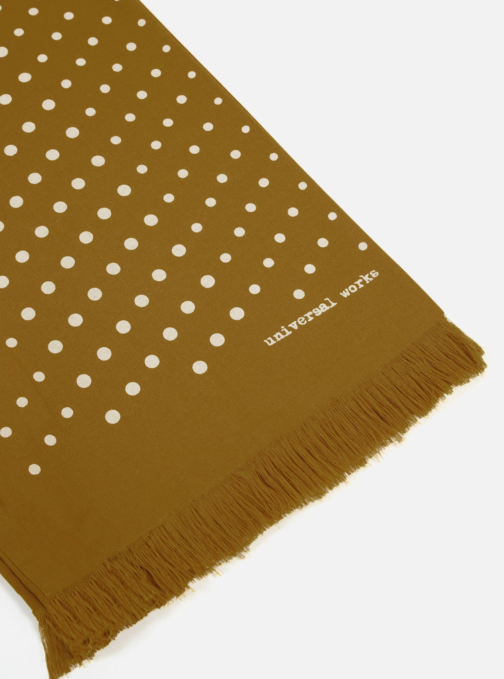 Close-up of the Mustard Dot Print Short Scarf on a white surface. The scarf is mustard-colored with white dots and a fringed edge. "universal works" is printed on the scarf.