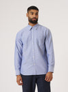 A man stands against a white backdrop, modeling the Blue Oxford Daybrook Shirt. The shirt is light blue, buttoned, and has a pocket. He wears dark pants.