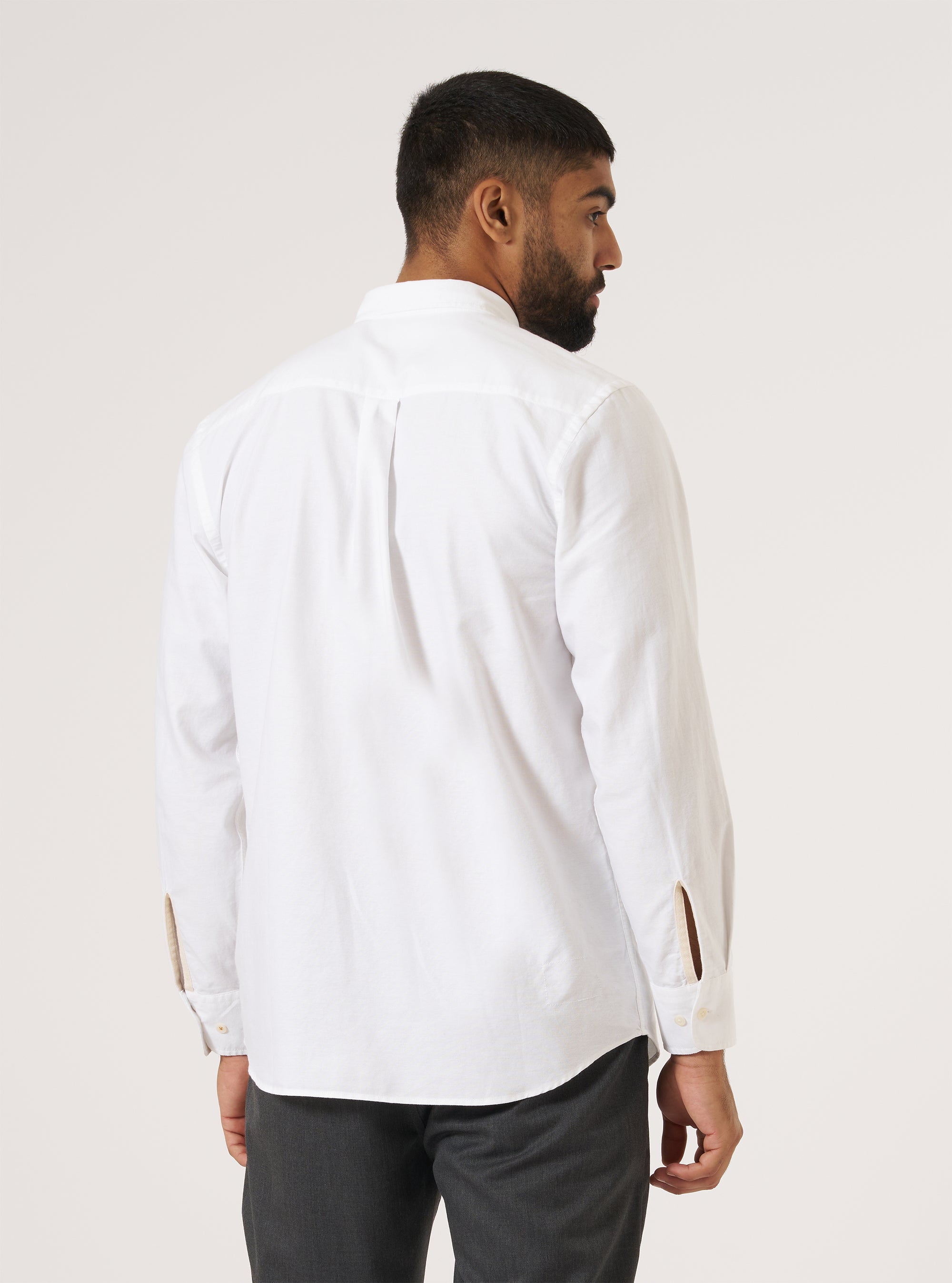 A man stands against a white backdrop, showcasing the back of a White Oxford Daybrook Shirt. The shirt is paired with gray pants, offering a clean, classic look.