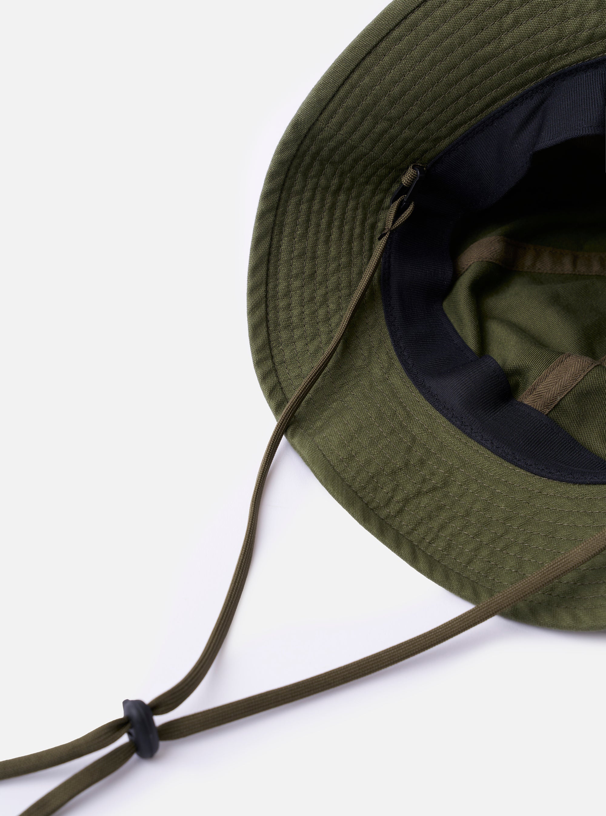 The cableami Dark Olive Baker Back Satin Cotton Metro Hat is shown as a flat lay against a plain white background. The image provides a detailed side-profile view of the hat, highlighting its six-panel construction, the slightly textured dark olive satin fabric, and the narrow, downward-sloping brim. A thin, dark adjustable chin strap with a plastic toggle is attached.