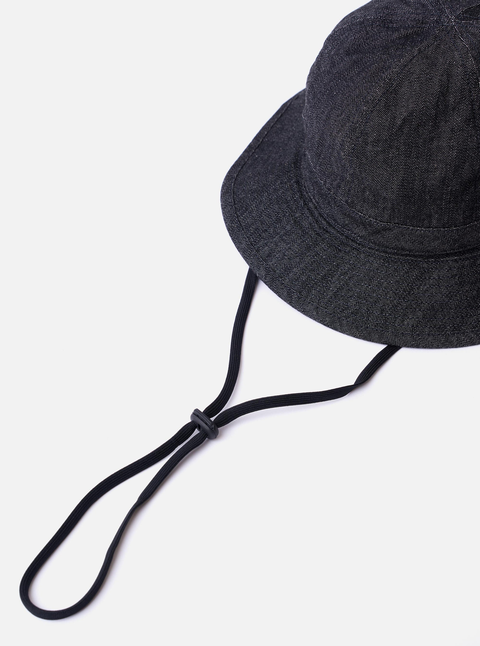 The cableami Black 10oz Denim Metro Hat is shown from a rear three-quarter angle as a flat lay against a plain white background. The image highlights the hat's rounded crown, the downward-sloping brim with tonal stitching, and the adjustable black strap cord with a toggle. The 10oz black denim fabric shows a subtle textured grain.