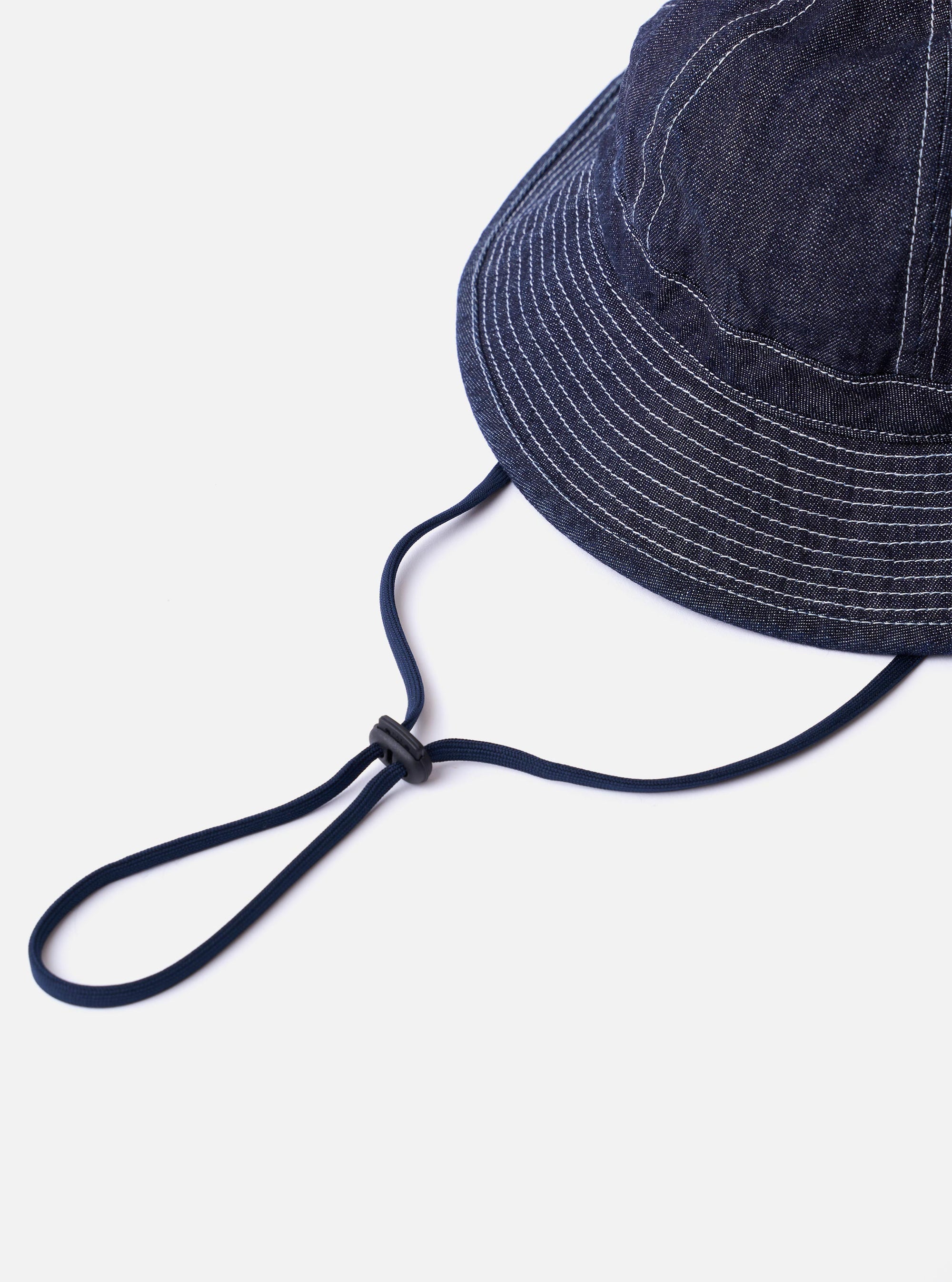 The cableami Indigo 10oz Denim Metro Hat is shown from a rear three-quarter angle as a flat lay against a plain white background. The image highlights the rounded crown and downward-sloping brim, as well as the adjustable drawcord and stopper at the back. The indigo denim fabric features a visible natural slub texture.