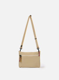 The master-piece x Universal Works Beige Canvas Sacoche is shown as a flat lay against a white background. The small rectangular shoulder bag is crafted from a durable beige canvas fabric and features a dual-branded logo patch on the front, a black adjustable shoulder strap with metal hardware, and a main compartment with a secure closure.