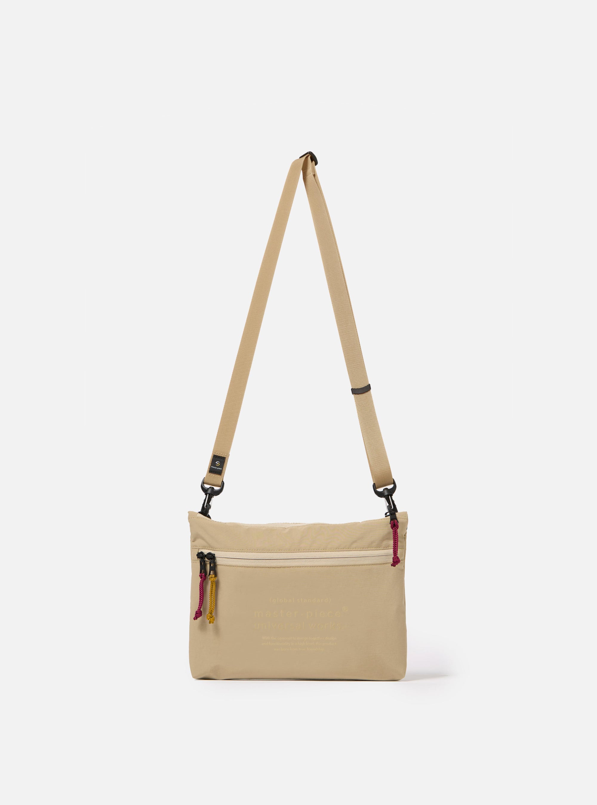 The master-piece x Universal Works Beige Canvas Sacoche is shown as a flat lay against a white background. The small rectangular shoulder bag is crafted from a durable beige canvas fabric and features a dual-branded logo patch on the front, a black adjustable shoulder strap with metal hardware, and a main compartment with a secure closure.