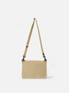 The master-piece x Universal Works Beige Canvas Sacoche is displayed from a side-angle as a flat lay against a plain white background. The image highlights the slim profile of the bag, the adjustable beige webbing shoulder strap with silver-tone metal hardware, and the durable canvas construction.