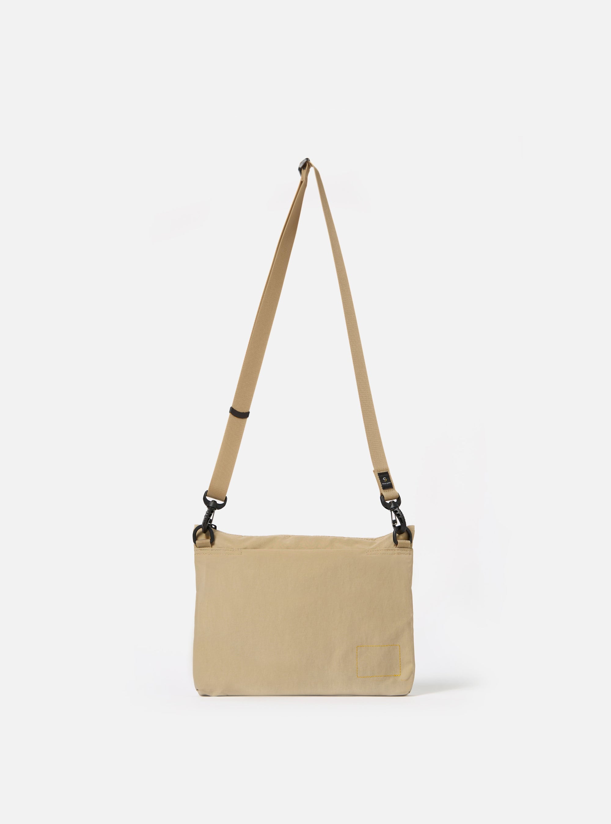 The master-piece x Universal Works Beige Canvas Sacoche is displayed from a side-angle as a flat lay against a plain white background. The image highlights the slim profile of the bag, the adjustable beige webbing shoulder strap with silver-tone metal hardware, and the durable canvas construction.