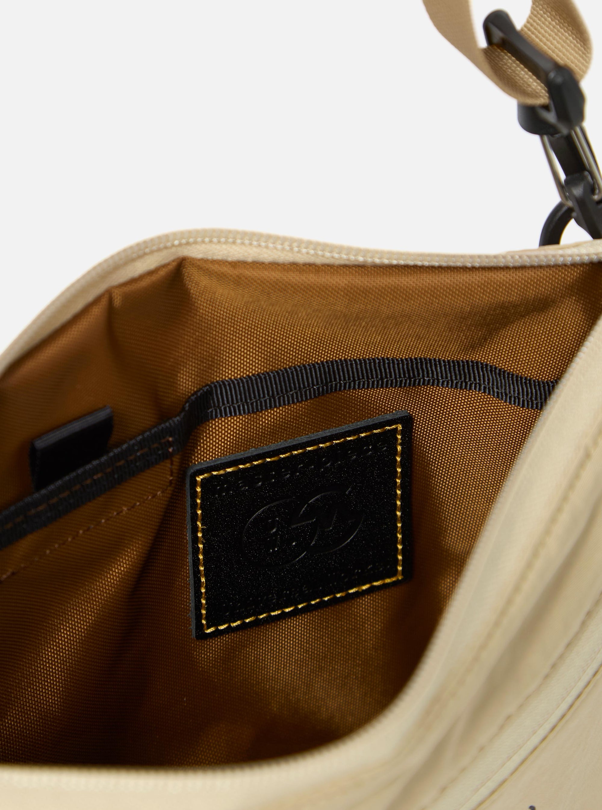 The master-piece x Universal Works Beige Canvas Sacoche is displayed from a rear angle as a flat lay against a plain white background. The image highlights the clean back panel of the bag, the adjustable black crossbody strap, and the silver-tone hardware. The beige canvas fabric has a visible, durable texture.