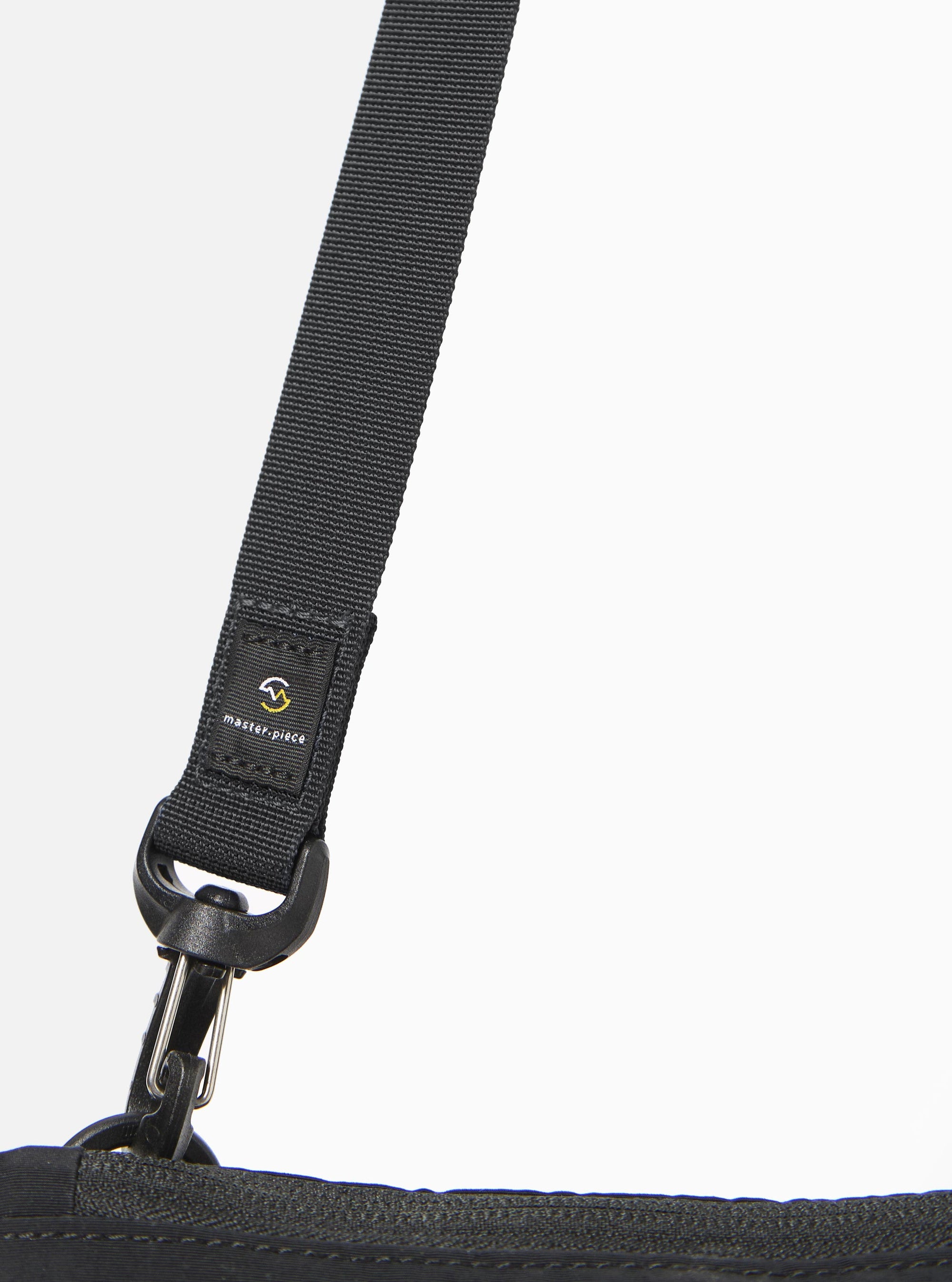 The master-piece x Universal Works Black Canvas Sacoche is shown from a rear three-quarter angle as a flat lay against a white background. The image highlights the adjustable black shoulder strap attached with metal clips, a rear slip pocket, and the durable texture of the black canvas fabric.