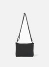 The master-piece x Universal Works Black Canvas Sacoche is shown from a rear angle as a flat lay against a white background. The image highlights the adjustable black shoulder strap attached with metal clips, a small brand tag near the bottom seam, and the clean, rectangular construction of the bag.