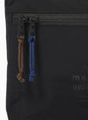 The master-piece x Universal Works Black Canvas Sacoche is displayed from a rear angle as a flat lay against a plain white background. The image highlights the adjustable black shoulder strap attached to the top corners and a small exterior pocket on the back panel. The black canvas fabric has a matte finish with visible tonal stitching.