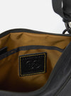 The master-piece x Universal Works Black Canvas Sacoche is shown in a detail view as a flat lay against a plain white background. The image focuses on the lower corner of the bag, highlighting the collaborative branding with a "master-piece" woven label and a printed "Universal Works" logo. The black canvas fabric has a fine, durable texture, and a portion of the adjustable shoulder strap is visible.