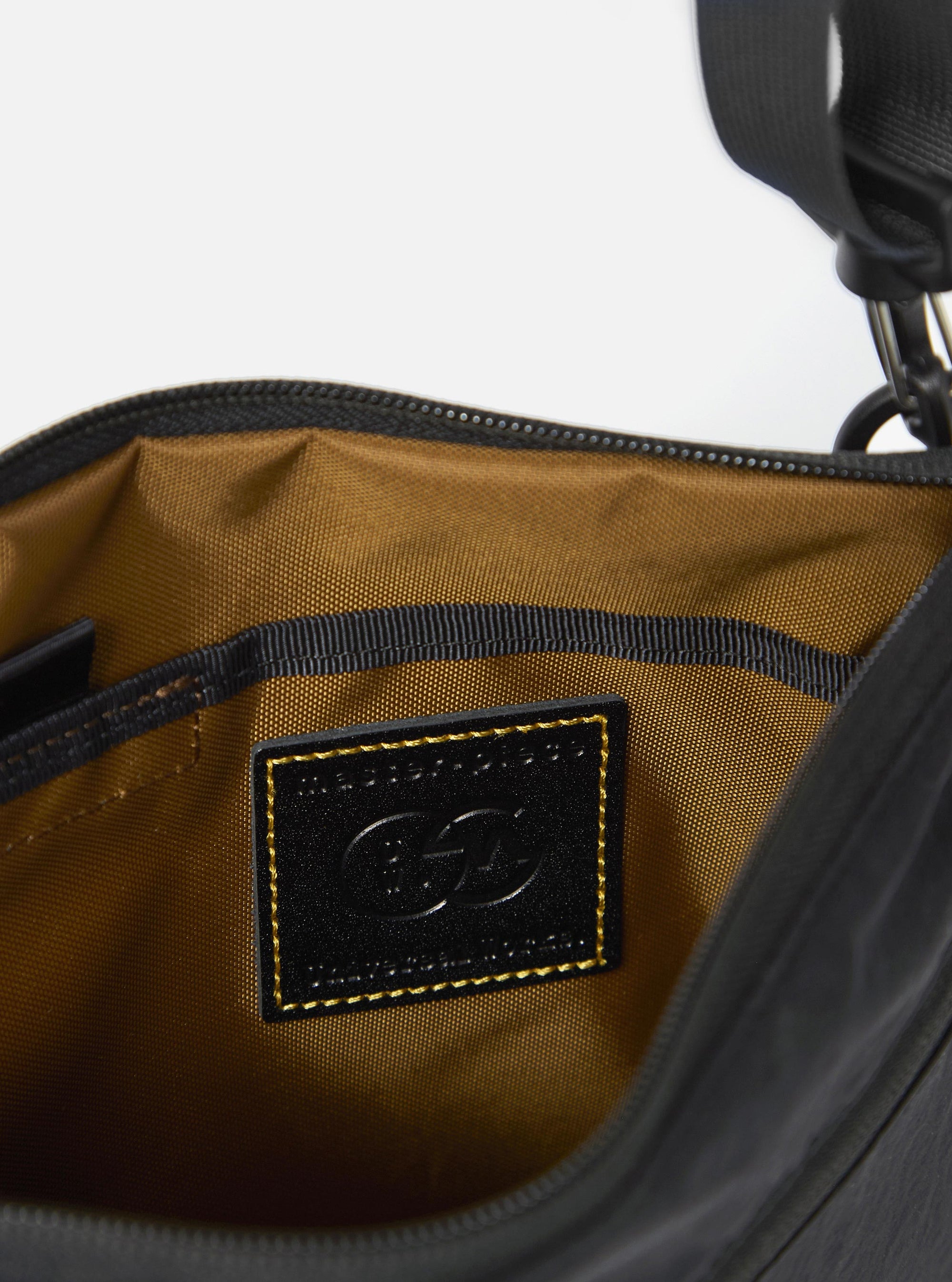 The master-piece x Universal Works Black Canvas Sacoche is shown in a detail view as a flat lay against a plain white background. The image focuses on the lower corner of the bag, highlighting the collaborative branding with a "master-piece" woven label and a printed "Universal Works" logo. The black canvas fabric has a fine, durable texture, and a portion of the adjustable shoulder strap is visible.