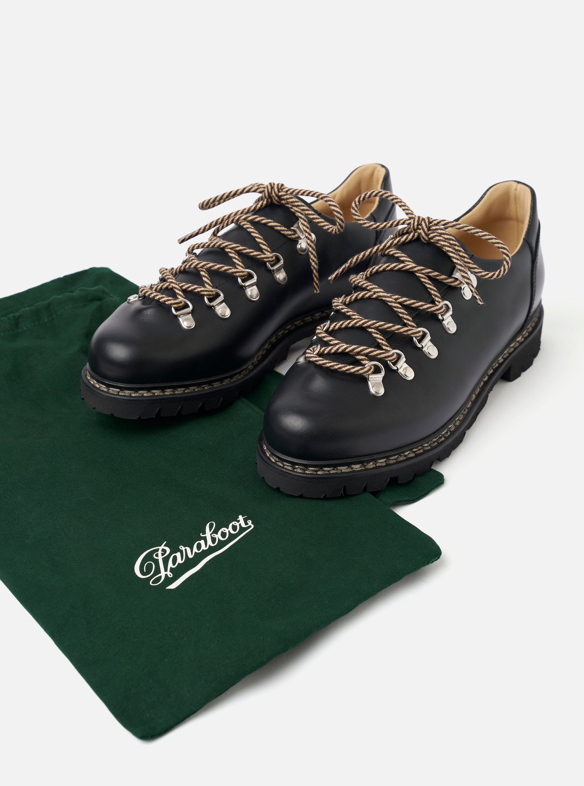 The Paraboot Black Leather Clusaz Jannu is shown in a studio shot against a plain white background. The low-top mountaineering shoe is crafted from smooth black leather and features metal D-ring eyelets with black rope laces. The design is characterized by a prominent Norwegian welt with visible stitching and a thick, rugged black rubber lug sole.