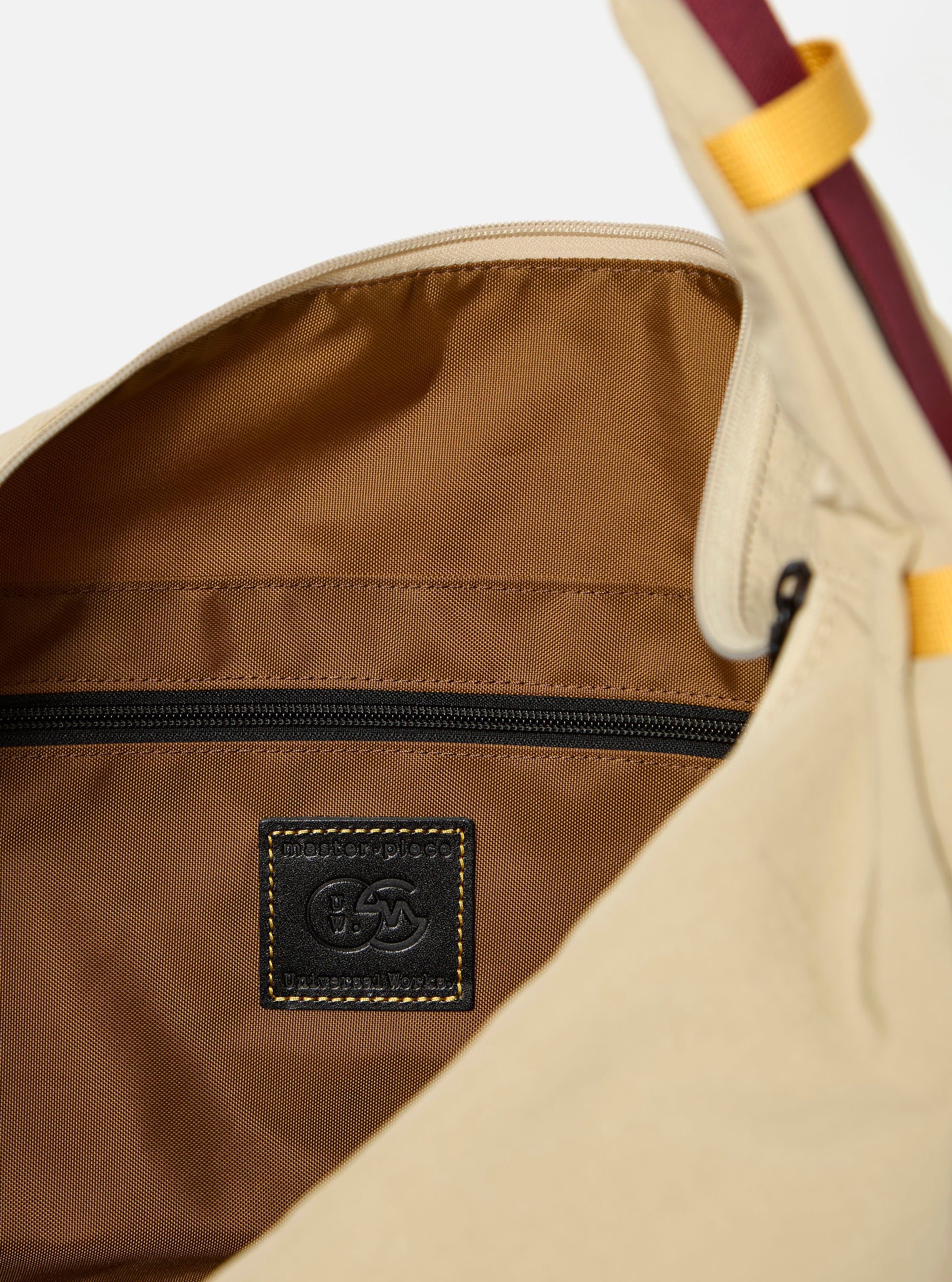 The master-piece x Universal Works Beige Large Canvas Shoulder Bag is shown from the back as a flat lay against a plain white background. The image highlights the wide, adjustable shoulder strap, a large external patch pocket with a dual-branded master-piece and Universal Works logo, and the durable beige canvas construction.
