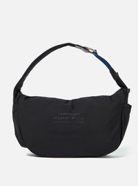 The master-piece x Universal Works Black Large Canvas Shoulder Bag is shown as a flat lay against a white background. The bag is made of black canvas and features two top carry handles, a detachable shoulder strap, and a front outer pocket with a visible dual-branded patch. It has a press stud fastening at the top and additional press stud detailing at the bottom.