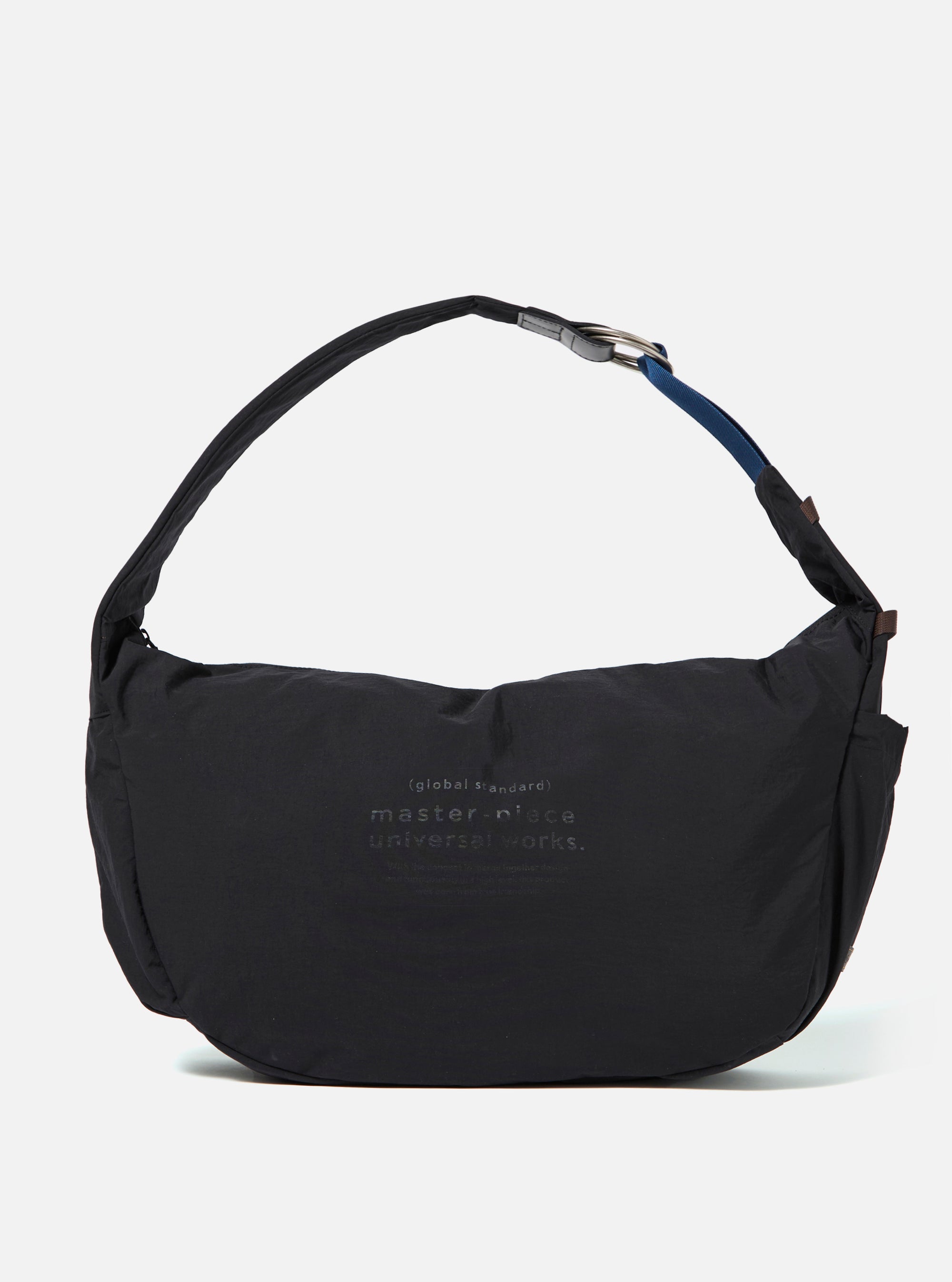 The master-piece x Universal Works Black Large Canvas Shoulder Bag is shown as a flat lay against a white background. The bag is made of black canvas and features two top carry handles, a detachable shoulder strap, and a front outer pocket with a visible dual-branded patch. It has a press stud fastening at the top and additional press stud detailing at the bottom.