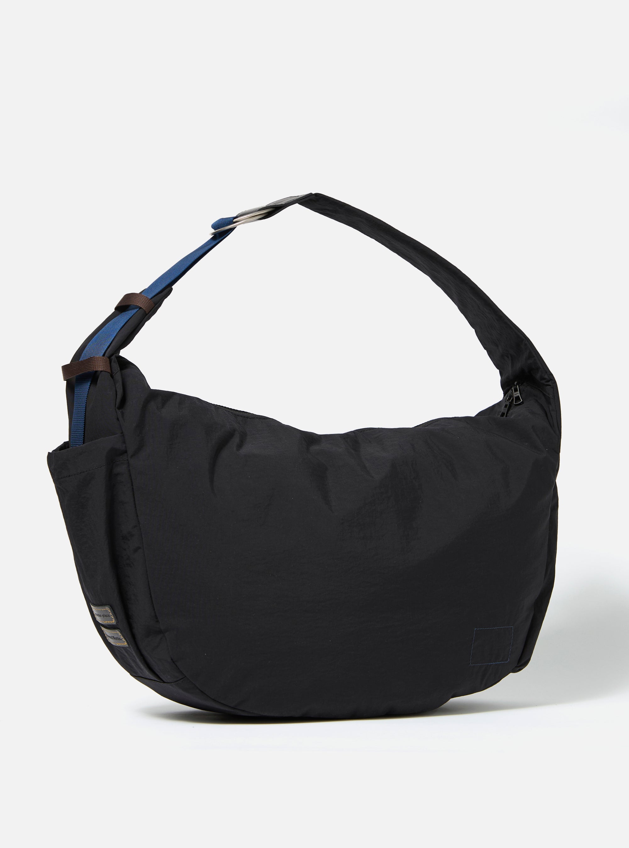 The master-piece x Universal Works Black Large Canvas Shoulder Bag is shown from a rear three-quarter angle as a flat lay against a plain white background. The image highlights the bag's structured rectangular shape, a small external pocket with a silver press stud, and a woven carry handle. The black canvas fabric has a smooth, matte finish.