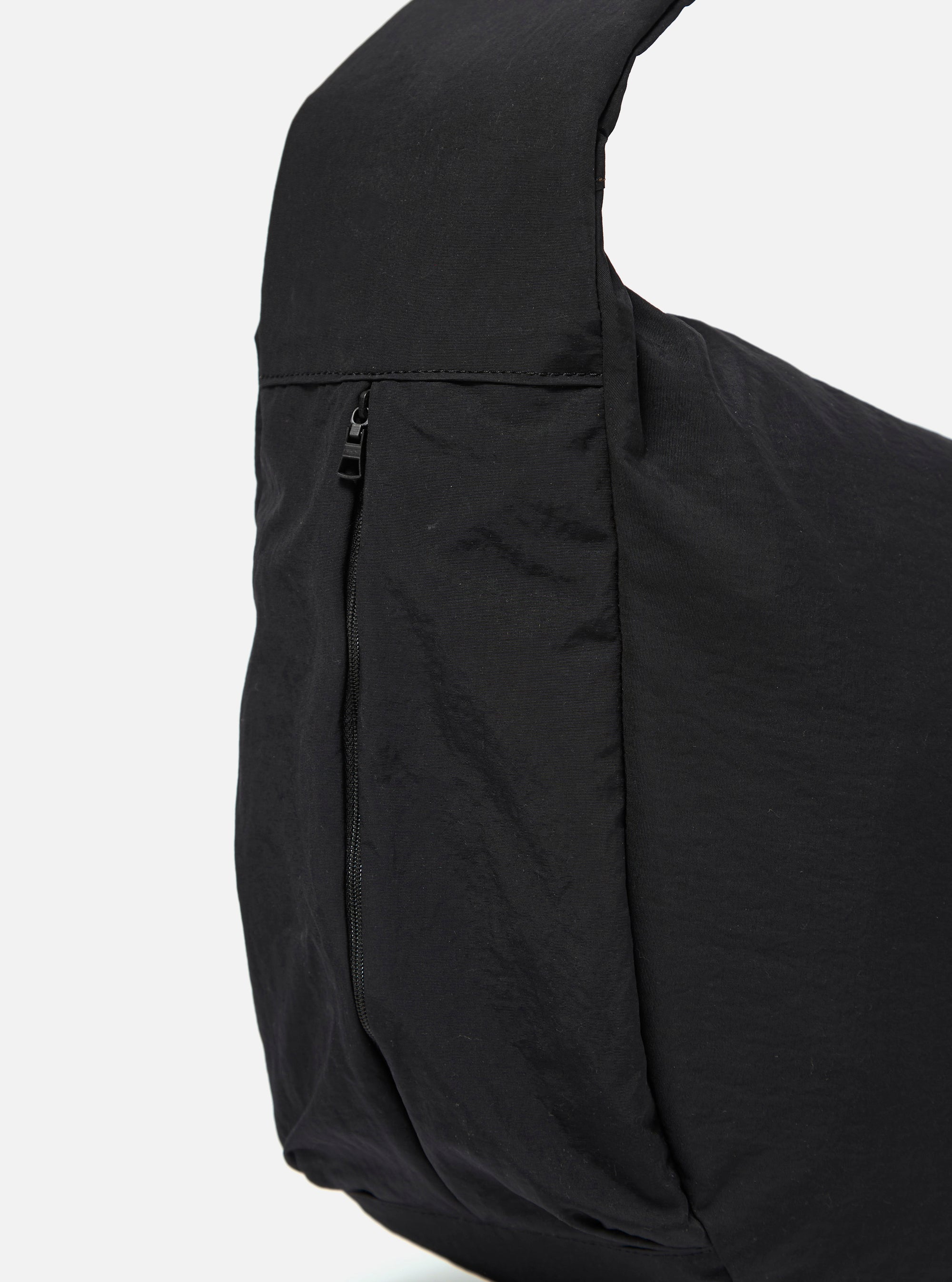 The master-piece x Universal Works Black Large Canvas Shoulder Bag is shown from the back as a flat lay against a plain white background. The image highlights the bag's durable black canvas construction, featuring a small woven brand tag near the bottom seam and clean tonal stitching throughout.