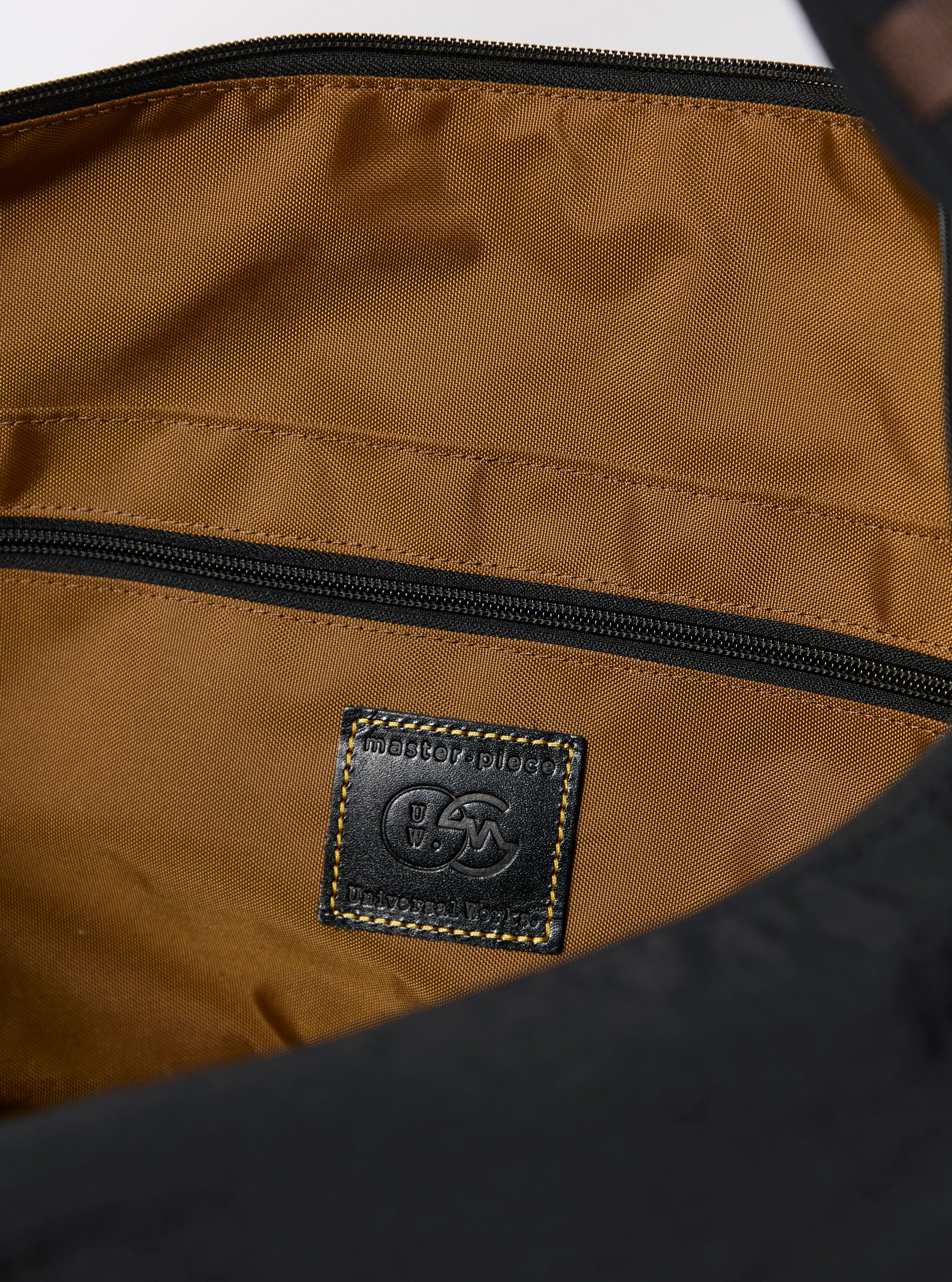 The master-piece x Universal Works Black Large Canvas Shoulder Bag is shown from a rear three-quarter angle as a flat lay against a plain white background. The image highlights the bag's large rectangular shape, the durable black canvas construction, and a side-entry zipper. A black adjustable shoulder strap is attached to the top corners, and a small collaborative brand tag is visible near the bottom edge.