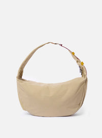 The master-piece x Universal Works Beige Medium Canvas Shoulder Bag is displayed against a plain white background. The bag features a rectangular silhouette with a main compartment, a front flap closure, and an adjustable shoulder strap made of webbing. A small collaborative brand patch is visible on the front. The beige canvas material has a clean, durable texture.