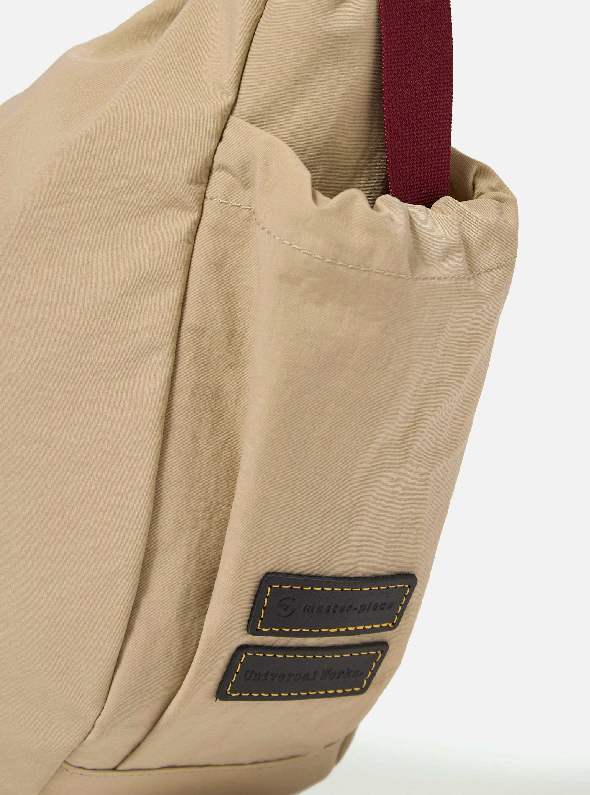 The master-piece x Universal Works Beige Medium Canvas Shoulder Bag is shown from the back as a flat lay against a plain white background. The image highlights the adjustable webbing cross-body strap, a small exterior slip pocket, and the tonal texture of the beige canvas fabric.