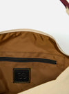 The master-piece x Universal Works Beige Medium Canvas Shoulder Bag is shown from the back as a flat lay against a plain white background. The image highlights the adjustable shoulder strap, a small exterior zippered pocket, and a rear sleeve for attaching to a suitcase handle. The bag is made from a durable beige canvas with a visible dual-branded patch.