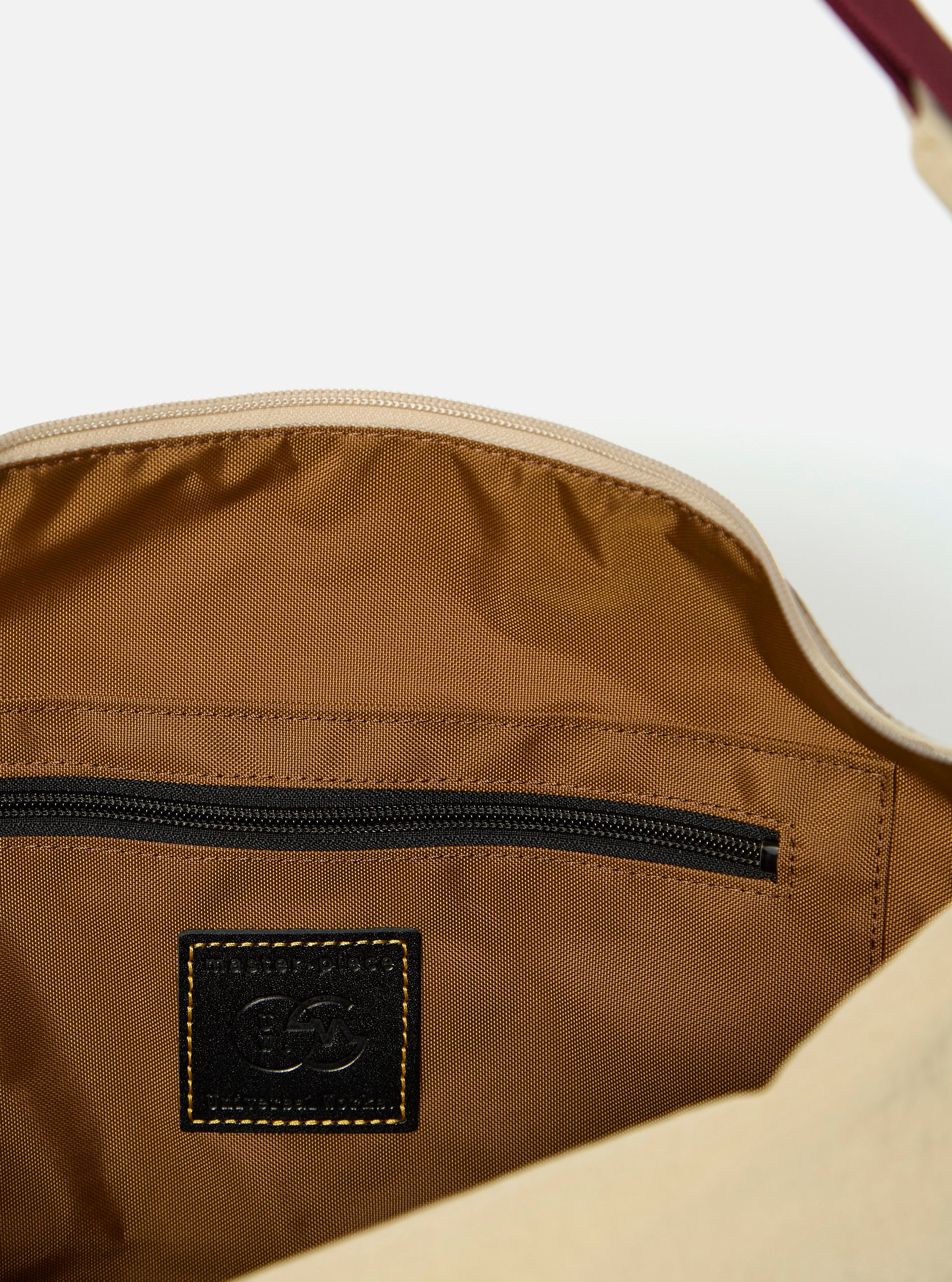 The master-piece x Universal Works Beige Medium Canvas Shoulder Bag is shown from the back as a flat lay against a plain white background. The image highlights the adjustable shoulder strap, a small exterior zippered pocket, and a rear sleeve for attaching to a suitcase handle. The bag is made from a durable beige canvas with a visible dual-branded patch.