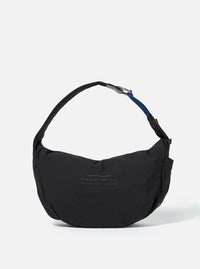 The master-piece x Universal Works Black Medium Canvas Shoulder Bag is shown as a flat lay against a white background. The bag is made of black canvas and features two top carry handles, a front patch pocket with a dual-branded logo patch, and silver-tone hardware including a detachable shoulder strap.
