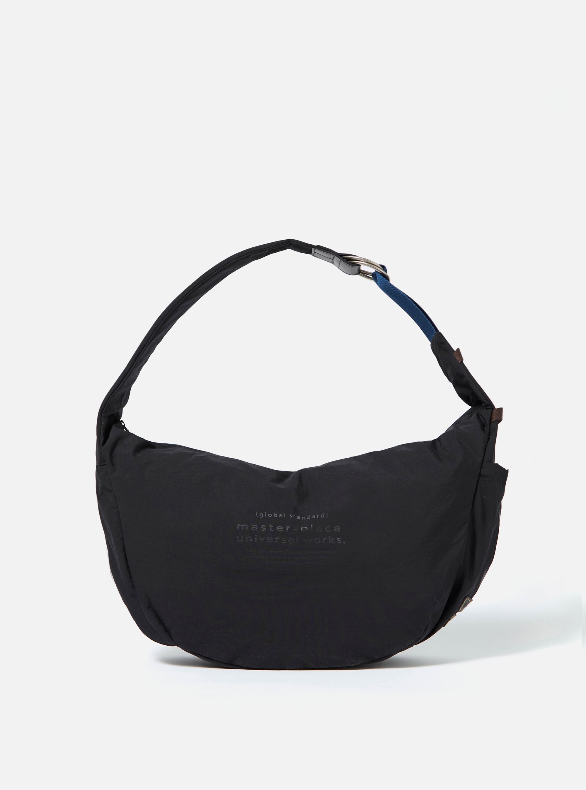 The master-piece x Universal Works Black Medium Canvas Shoulder Bag is shown as a flat lay against a white background. The bag is made of black canvas and features two top carry handles, a front patch pocket with a dual-branded logo patch, and silver-tone hardware including a detachable shoulder strap.