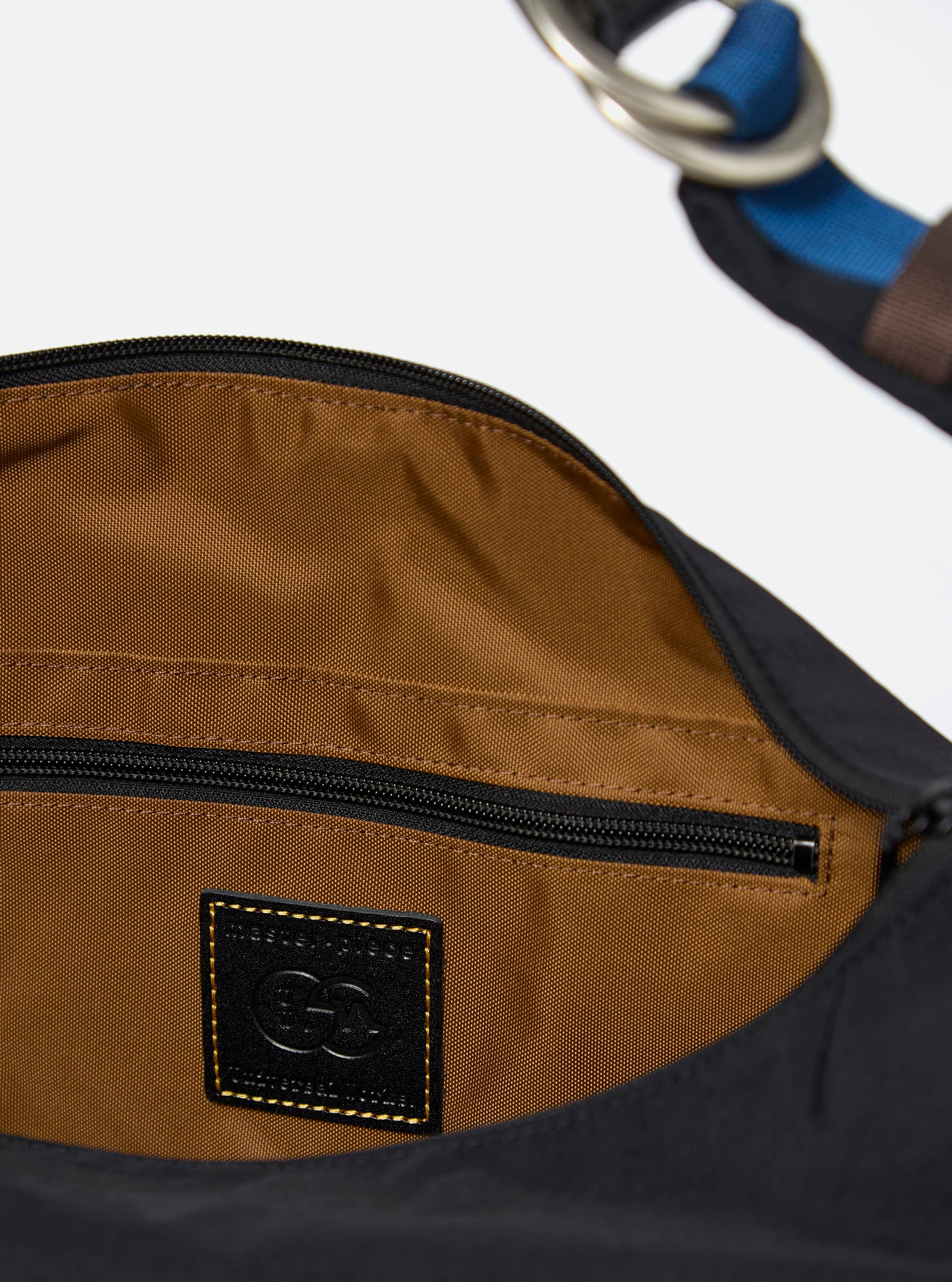 The master-piece x Universal Works Black Medium Canvas Shoulder Bag is shown from the back as a flat lay against a plain white background. The image highlights the clean, durable black canvas construction, featuring a horizontal strap across the center for attaching to a suitcase handle. A small, dual-branded logo patch is visible near the bottom edge.