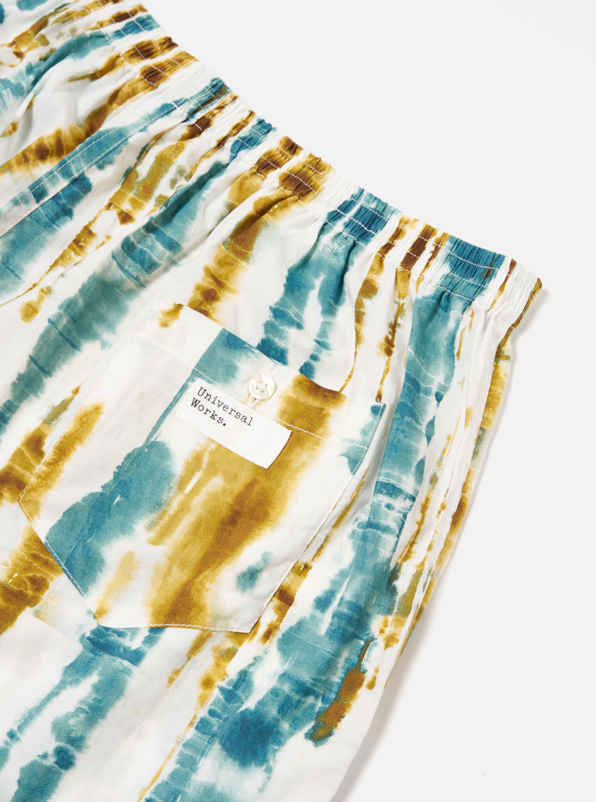 Close-up of the Ecru Paint Print Boxer Short on a white surface. Features a blue and brown tie-dye pattern, elastic waist, and a pocket with a button. 