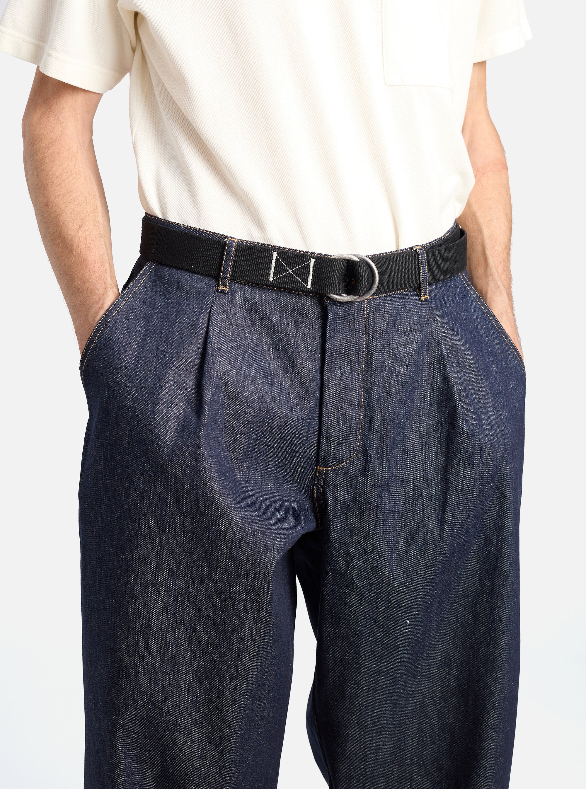 A model wears a Black Canvas O Ring Belt with dark denim pants and a cream t-shirt. The background is a clean white. The belt is black canvas with a silver O ring.