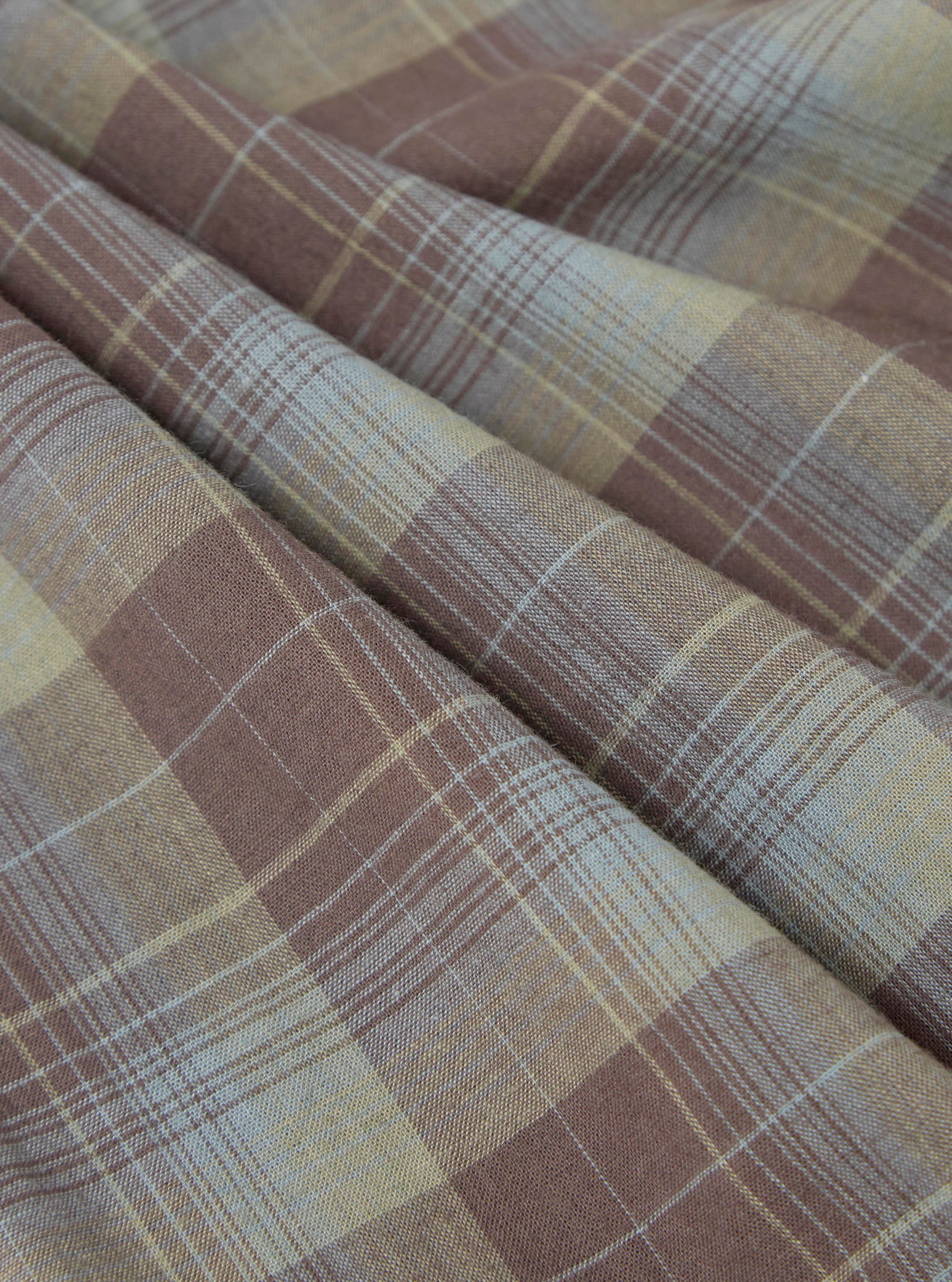 Close-up of the Green/Brown Check Pyjama Pant fabric. The material is folded, showing a detailed plaid pattern in brown, green, and white. The background is a plain surface.