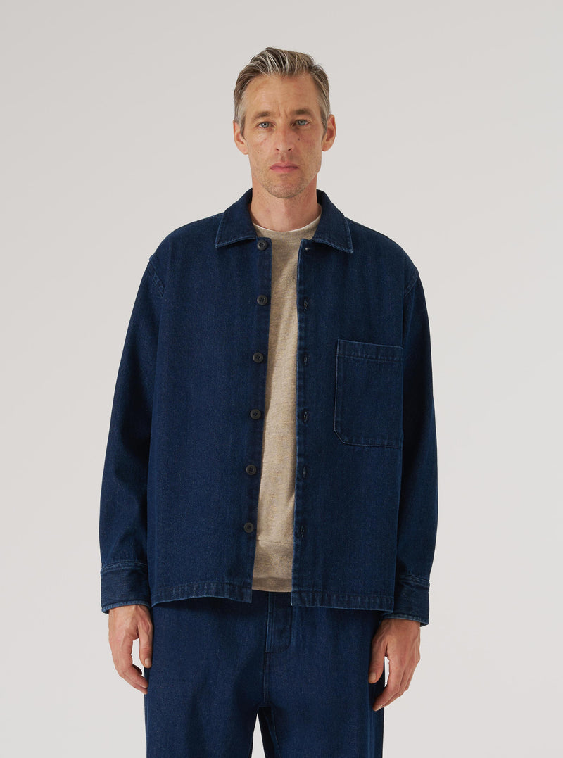 A man models the Indigo Work Denim Fatigue Shirt in a studio setting. The shirt is dark blue denim with a relaxed fit. He wears a beige t-shirt underneath. White backdrop.