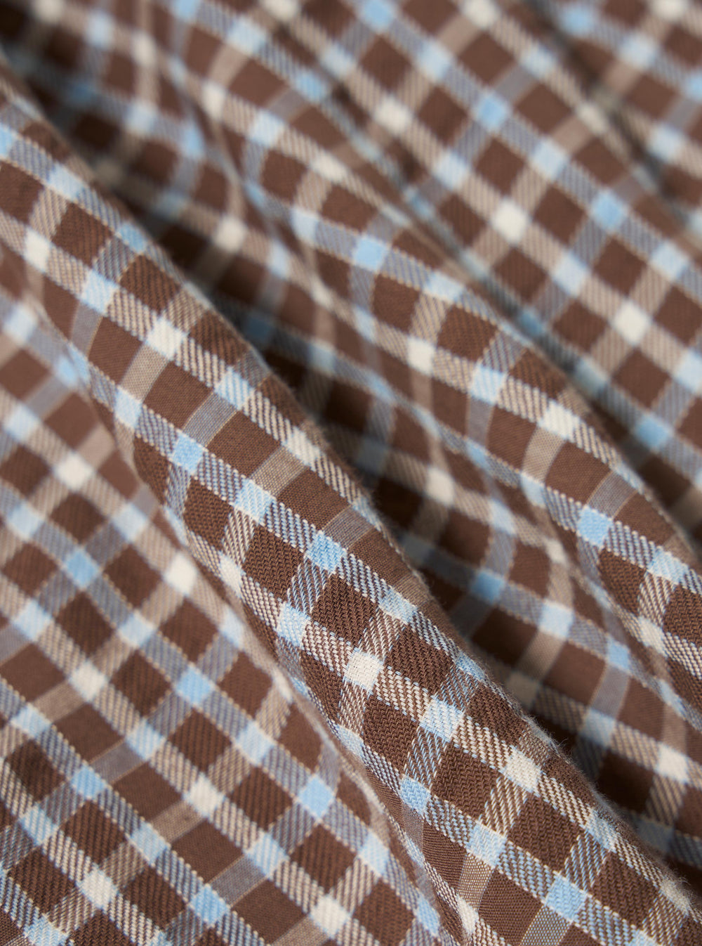 Close-up of the Brown Madison Check Station Shirt fabric. Features a brown, light blue, and white check pattern. The material is folded on a flat surface.