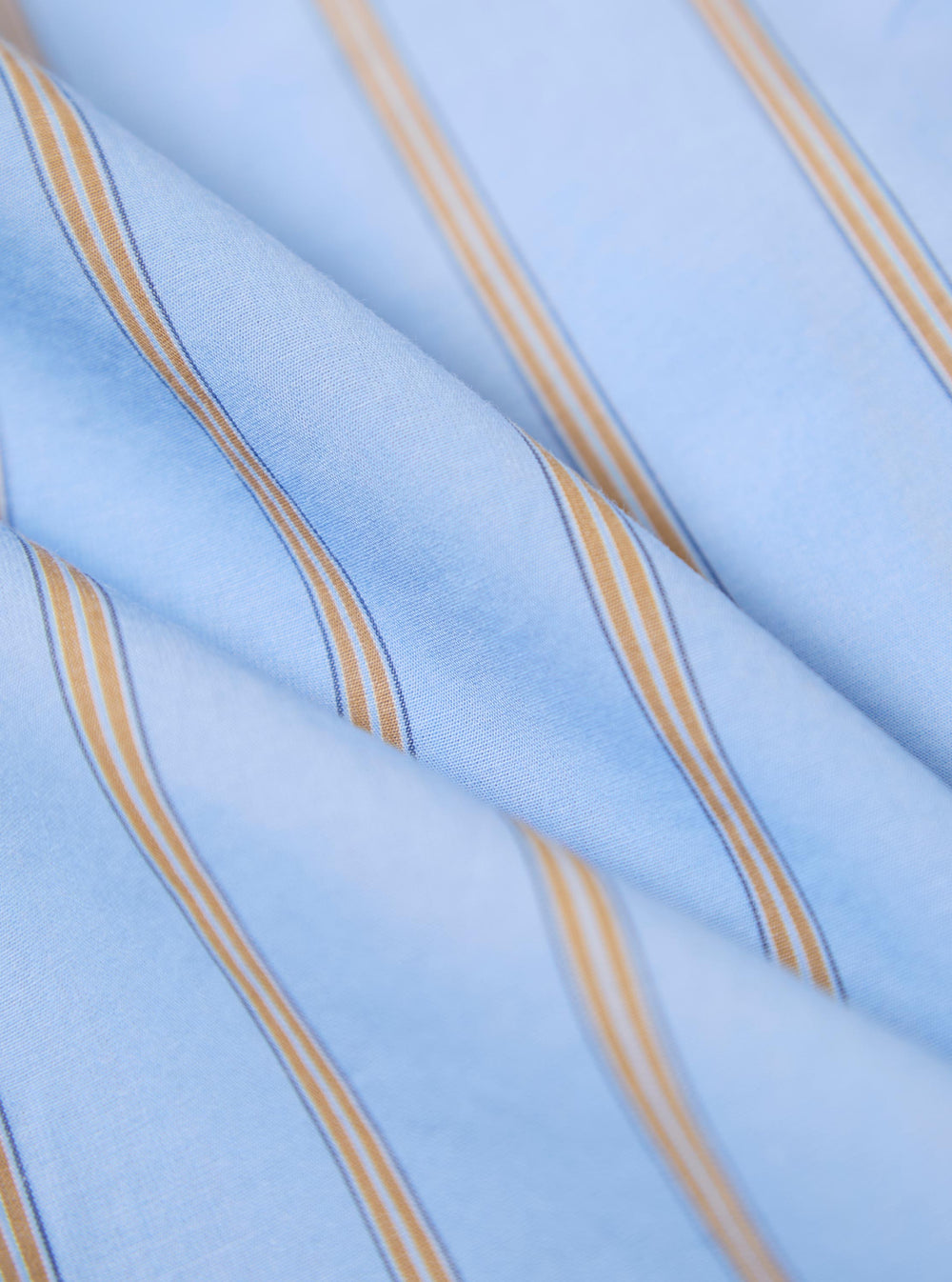 Close-up of the Blue Pop Stripe Four Lazy Day Shirt fabric. Light blue with thin gold and blue stripes. The shirt is folded on a white surface.