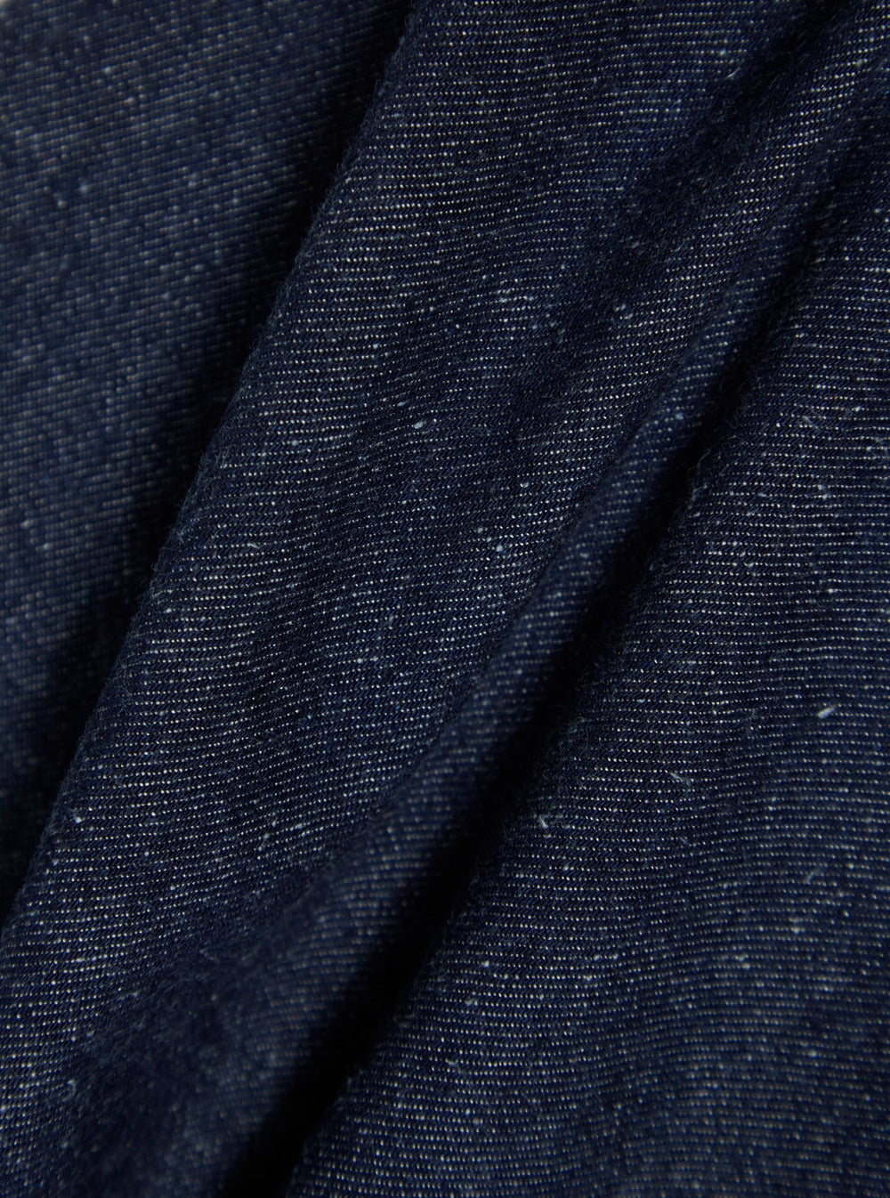 Close-up of the Indigo Future Denim Oxford Pant fabric. The dark blue denim is folded on a plain surface, showing the texture and weave.