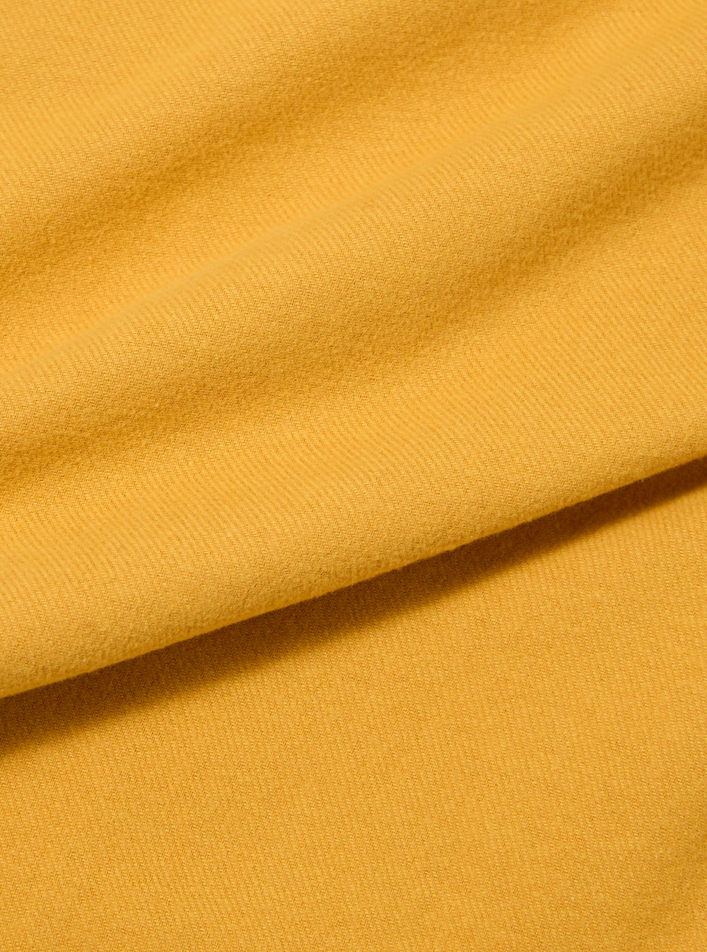 Close-up of the Gold Alaska Cotton Utility Shirt fabric, showing its texture and color. The shirt is laid out on a flat surface, highlighting the material's quality.