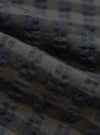 Close-up of the Charcoal/Navy Seersucker Plaid Pyjama Pant fabric. The material shows a textured plaid pattern in charcoal and navy. The fabric is folded, highlighting its soft texture.