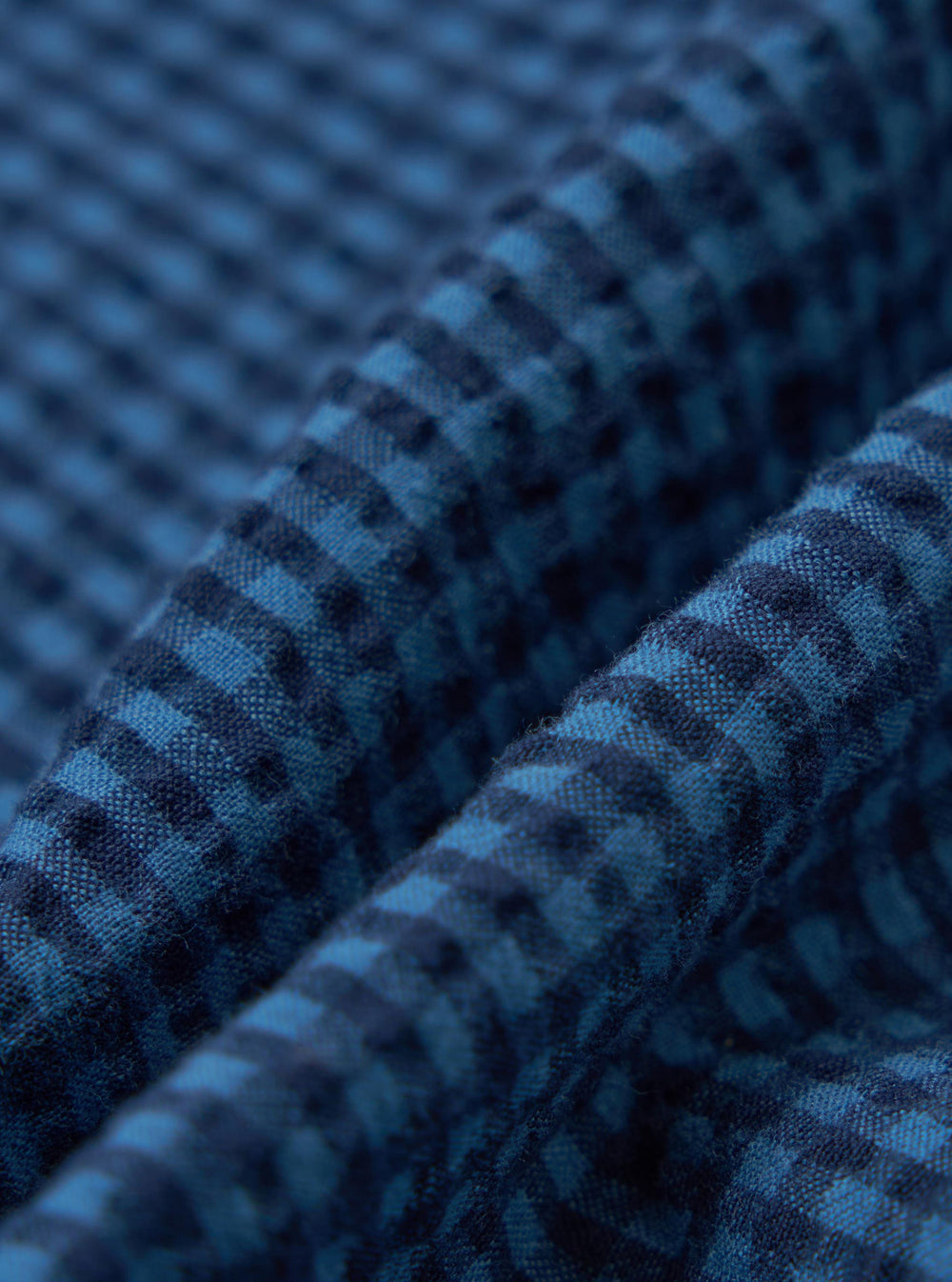 Close-up of the Blue Gingham Seersucker Pyjama Short fabric. The material is folded, showing the texture and pattern on a plain background.