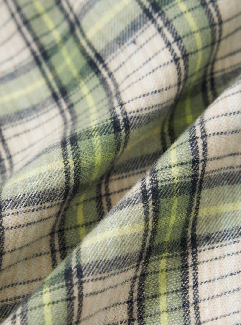 Close-up of the Green Busy Plaid Short Pyjama Set fabric. The soft cotton blend features a green, yellow, and black plaid pattern. The material is folded on a neutral surface.