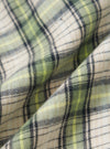Close-up of the Green Busy Plaid Short Pyjama Set fabric. The soft cotton blend features a green, yellow, and black plaid pattern. The material is folded on a neutral surface.