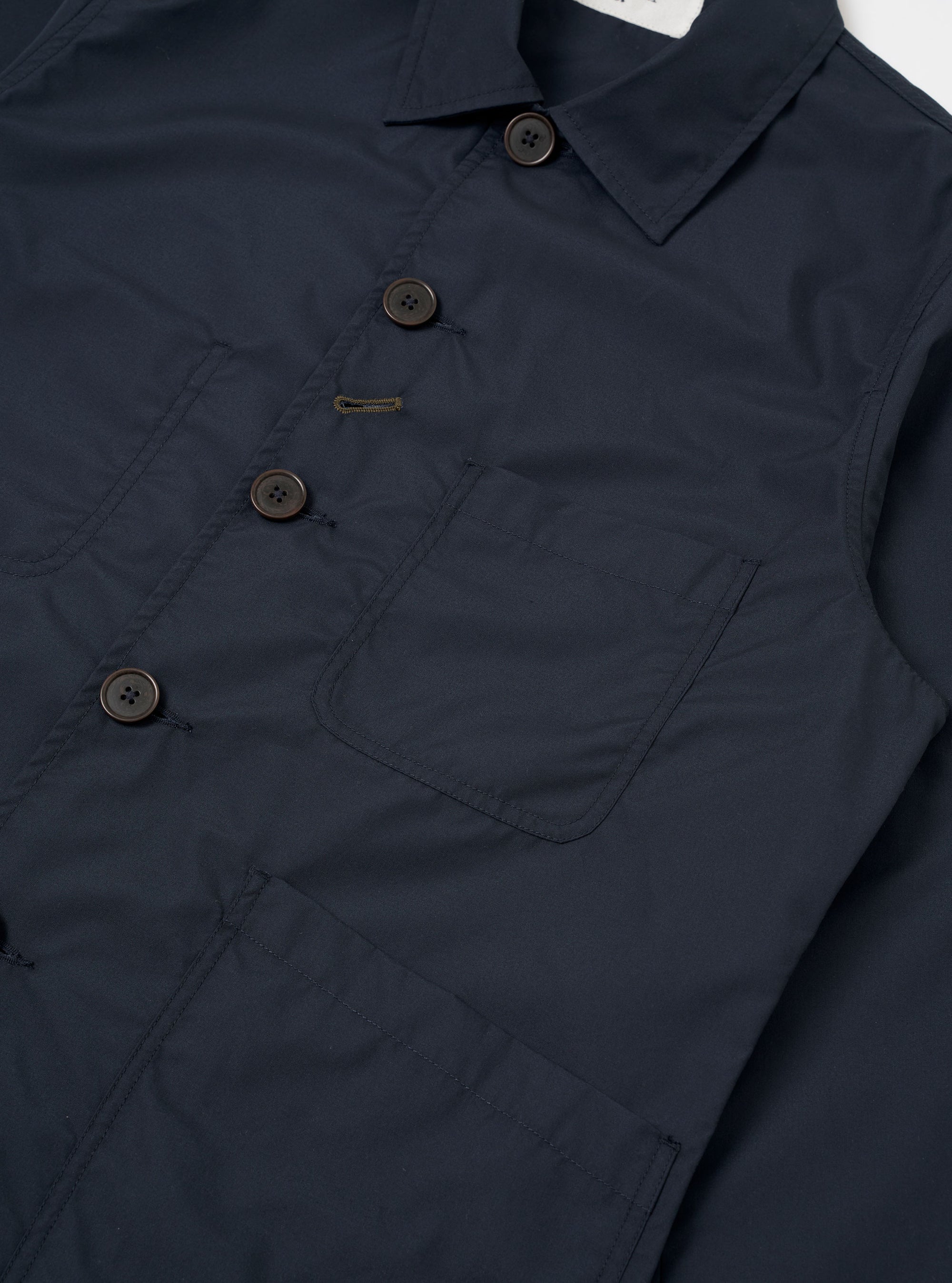The Navy Recycled Polytech Bakers Jacket is shown as a flat lay against a white background. This chore-style jacket features a five-button front closure, a classic pointed collar, and a curved front hem. It has one patch pocket on the chest and two larger patch pockets at the hips. The fabric is a smooth, dark navy navy blend of recycled polyester and cotton.