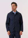 A model wears the Navy Recycled Polytech Bakers Jacket over a white shirt and patterned tie against a plain white background. The jacket is a deep navy blue with a regular fit, featuring a five-button front closure, a classic pointed collar, and a curved front hem. Prominent details include two large patch pockets at the waist and one smaller patch pocket on the left chest. The model is shown from the waist up.