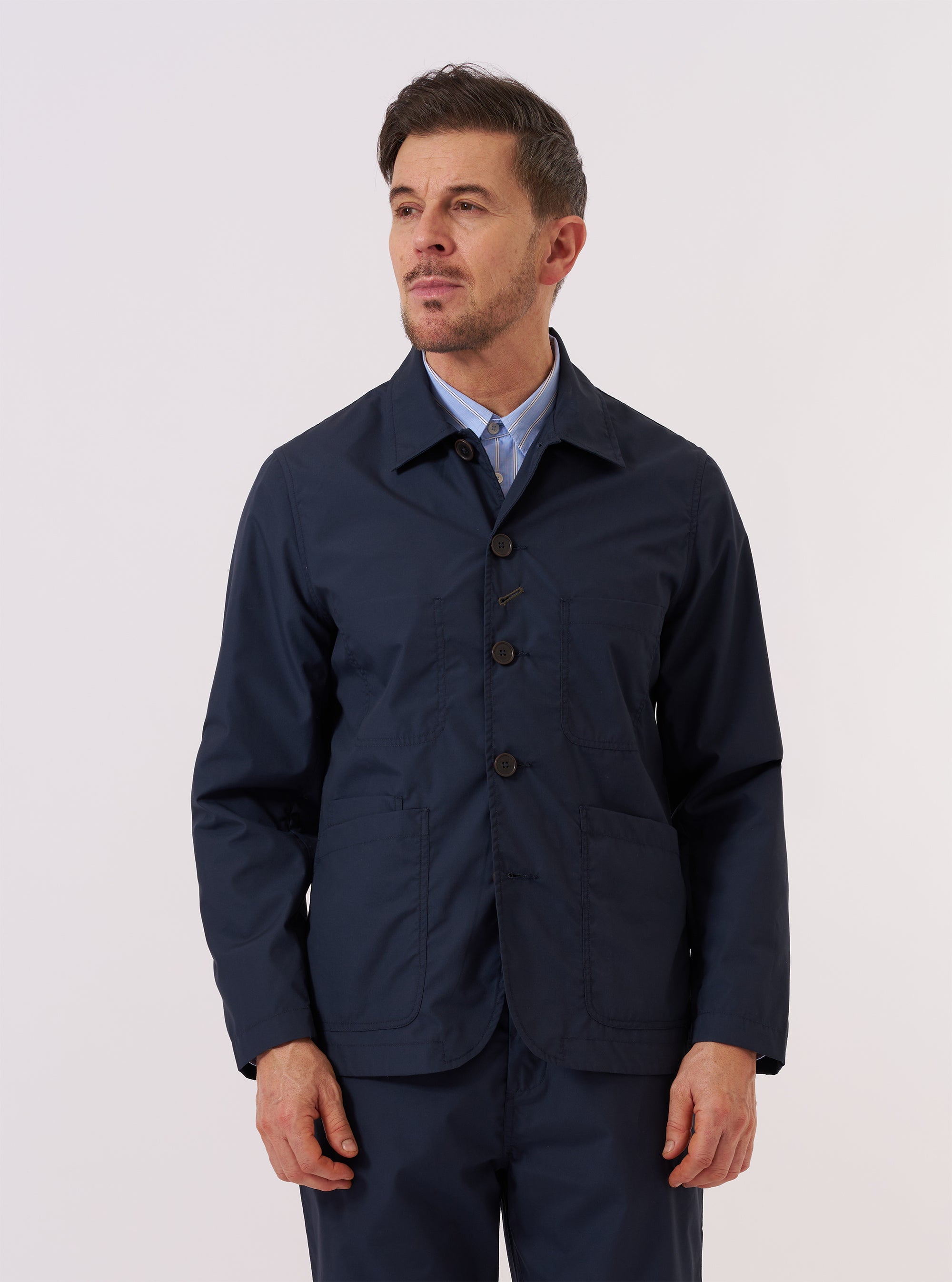 A model wears the Navy Recycled Polytech Bakers Jacket over a white shirt and patterned tie against a plain white background. The jacket is a deep navy blue with a regular fit, featuring a five-button front closure, a classic pointed collar, and a curved front hem. Prominent details include two large patch pockets at the waist and one smaller patch pocket on the left chest. The model is shown from the waist up.