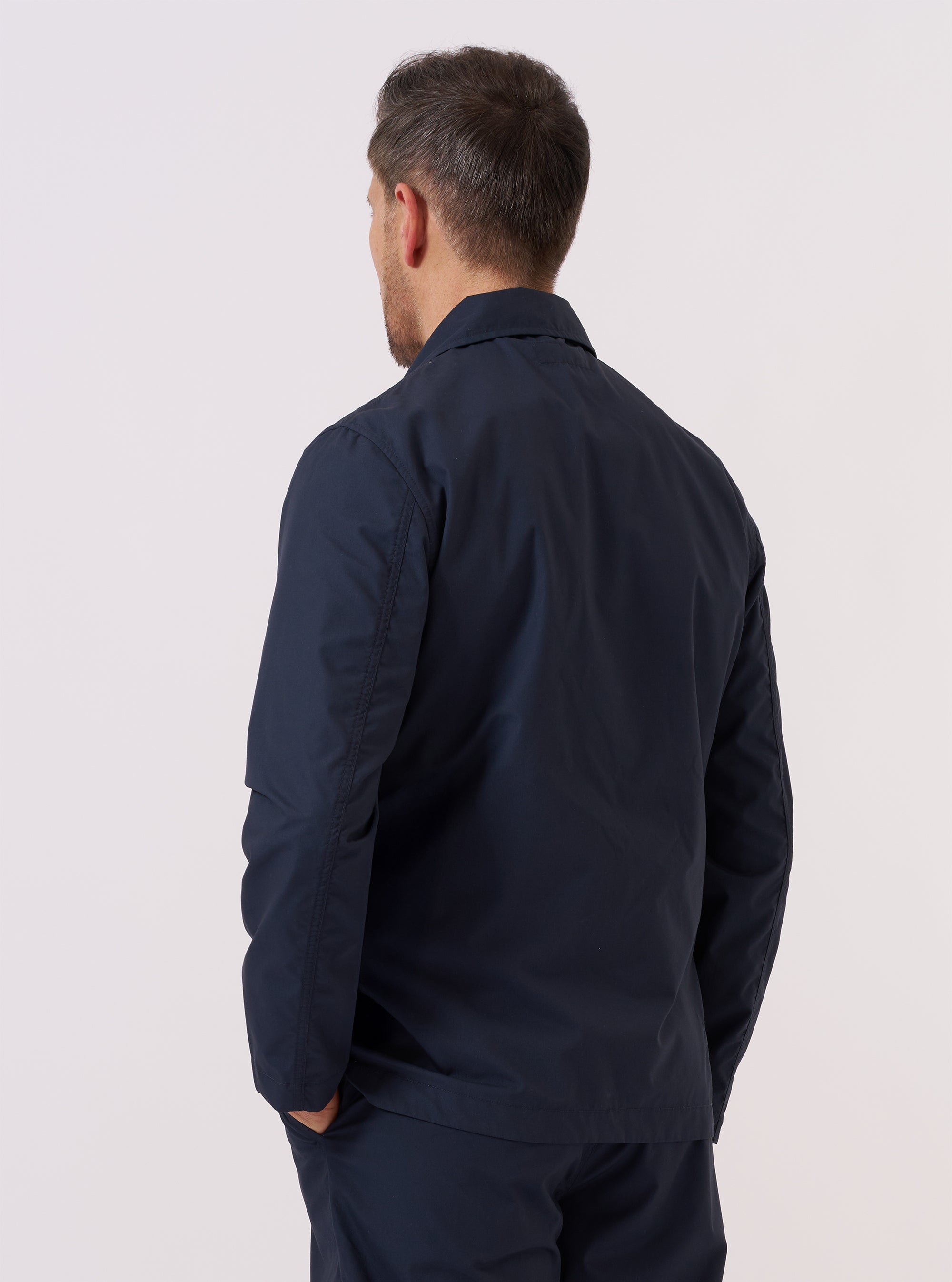 A model wears the Navy Recycled Polytech Bakers Jacket over a light-colored shirt, standing against a plain white background. The jacket is shown from a three-quarter angle, highlighting its classic collar, five-button front closure, and multiple large patch pockets on the front.